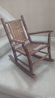 Front and side view of a vintage children's rocking chair with wooden frame and woven wicker seat, showing wear on wicker.