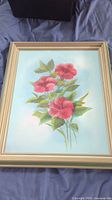 Full view of framed canvas artwork featuring pink flowers and green leaves on a blue background, placed flat on fabric.