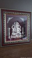 Framed 3D embossed silver Ganesh artwork with decorative frame and fabric background.