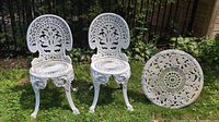 Pair of white cast aluminum patio chairs with intricate leaf and floral openwork designs on backrests and circular openwork seats, one chair missing bottom seat piece placed to the side.