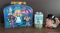 Full view of Alice in Wonderland doll set in plastic case, Funko Soda Mad Hatter can, and Royal Doulton Mad Hatter jug