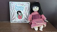 Japanese vinyl doll seated next to Once Upon a World Snow White hardcover book showing front covers and doll details