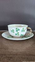 Side view of cream colored porcelain teacup and matching saucer with green shamrocks and bamboo style handle