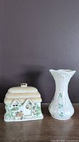 Front view of Lenox Irish Blessing trinket box shaped as a cottage with green vine decoration and gold chimney next to the Belleek Shamrock vase, both on a wooden surface against dark background