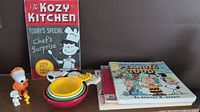 Three Peanuts books stacked neatly including Peanuts 2000 with Charlie Brown and Snoopy on cover, colorful Hallmark Peanuts measuring cups, Hallmark Kozy Kitchen metal sign with Lucy character, wind-up Snoopy toy, and two small Peanuts figures on brown surface.