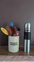 Ceramic kitchen utensil holder with wooden tongs, happy face spatula, blue spatula, and Finnish cheese slicer, all inside, next to Arcosteel stainless steel thermos.