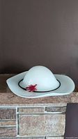 Front angled photo of large white Murano glass hat sculpture with black band and red flower detail on stone surface against dark wall