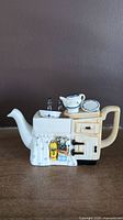 Front view of ceramic teapot shaped like kitchen sink and stove with miniature cleaning products on towel rail.