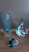 Photo showing the three glass pieces on a wooden surface: tall rectangular blue and clear glass vase, a colorful signed Julia glass heart paperweight, and a blue Robert Held glass heart paperweight.