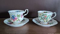Pair of Royal Albert teacups and saucers displayed side by side on a wooden surface against a brown background. One cup with Summertime floral design and one with Holly design.