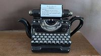 Full front view of black ceramic teapot shaped like a vintage typewriter with detailed keys and metallic carriage.