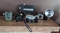 Five vintage cameras including a green Dacora box camera, Kodak Baby Brownie bakelite black camera, Kodak Brownie Holiday Flash with flash, Bell & Howell Autoload camera, and Bell & Howell 8429 Super 8 with carry bag and instructions.