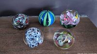 Five vintage glass paperweights displayed together on a surface, showing various colors and swirl patterns.