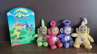 Four plush Teletubbies toys (green, pink, purple, yellow) displayed upright in a row with the Teletubbies Like to Dance book standing behind them.