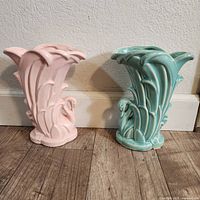 Both pink and turquoise ceramic swan vases shown side by side on floor with wall background.