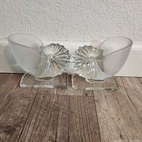 Front view of two frosted art glass nautilus seashells placed side by side on wooden floor against a white wall.