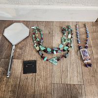 Art Deco style hand mirror with hexagonal reflective surface and silver-tone handle alongside two beaded multi-stone necklaces and silver earrings on card.