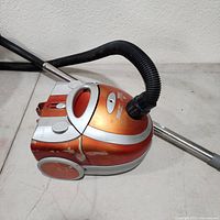 Bissell Zing vacuum cleaner side view showing orange and silver body with hose and metal wand attached.