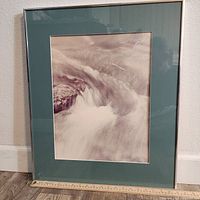 Front view of the framed monochrome art print 'Bubbling Brook' by William Plante with light green matte and silver frame, resting against a baseboard with ruler for scale.
