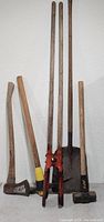 Full view of six large hand tools leaning against a wall including an axe, shovel, mattock, pickaxe, and mallet, showing wooden handles and metal heads.