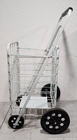 Side view of white metal rolling shopping cart showing wire basket, frame, and four wheels including large rear black rubber wheels.