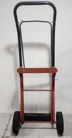 Front and second angle view of a metal and wooden moving furniture dolly standing upright, showing the black metal frame, reddish wooden support, wheels, and base plate.
