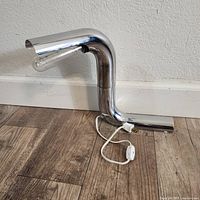 Chrome finished mid-century modern table lamp shown lying on wood floor near beige wall. Lamp arm is swiveling and holds an exposed tubular bulb.