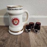 Full lot photo showing the large USC 1880 white ceramic mug next to four purple shot glasses with silver holders