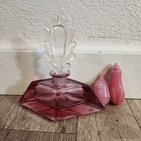 Front view showing purple perfume bottle with clear glass stopper alongside two small pink Peking snuff bottles of different sizes and shapes, all on wooden floor against white wall.