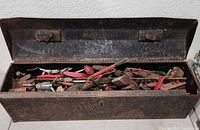 Open vintage metal toolbox showing interior filled with various hand tools such as pliers, wrenches, hammer, and screwdrivers, all showing signs of heavy use and rust.