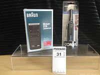 Braun 278 shaver original boxed product next to packaged Conair nose and ear hair trimmer placed on clear acrylic stand on striped fabric.