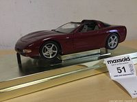 Front side view of maroon Corvette model car on a metal base, showing front details and wheel design.