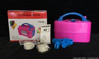 Photo showing the pink electric balloon pump, original packaging box with colorful balloons on front, and included balloon clips and tape accessories arranged in front of the pump.