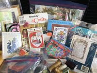 Photo showing assorted embroidery and needlework kits and patterns, including Simplicity Crafts and Jiffy kits, loose threads in plastic bags, and packaged fabric canvas.