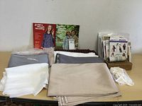Assorted fabrics in beige, grey, white, and off-white colors spread on a table with vintage buttonholer and sewing patterns in the background.