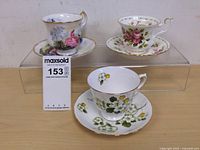 Three tea cups on matching saucers displayed on a clear acrylic riser on a wooden surface, showing floral designs and gold accents.