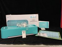 Turquoise LOKLIK cutting machine with power adapter and box in the background, accompanied by cutting board and manuals.