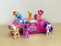 Six My Little Pony figurines displayed on pink branded My Little Pony box. One figurine (Twilight Sparkle) shows a broken tail.