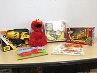 Lot showing all items including Elmo plush, CAT Power Hauler truck in box, WOW Roll-it Riley toy box, Clifford book, and puzzle sheet on a table.