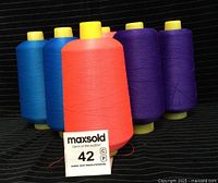Six large spools of serger thread in blue, coral, and purple colors, each wound around a spool with a yellow plastic base, displayed on a black textured surface.