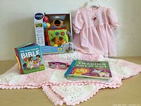 Full view of items including Vtech Sensory Cube, pink dress, pink crocheted blanket, My First Handy Bible book, and pink sunglasses