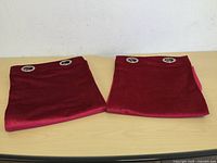 Two folded red velvet-like curtain panels lying flat on a surface, showing the fabric and grommet placement.