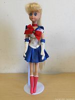 Front view of the 1996 Sailor Moon doll standing on the included white doll stand, showing blonde hair in ponytail, detailed sailor outfit with red bow and tall pink boots.
