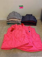 Photo showing stacks of vests in gray, beige, turquoise, and navy colors in the background, with 3 bright pink vests in the foreground.