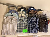 Overview of 20 men's dress shirts, assorted colors and patterns, mostly plaid and striped, folded and some bagged.