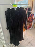 Two black velvet maxi dresses hanging on a rack, showing full length and overall styling including fringe sleeves and high tied necks.