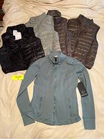 Five clothing pieces consisting of four down vests in dark and neutral colors and one blue gray zip-up jacket laid out on a bed.