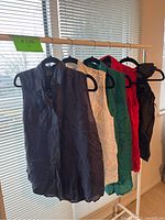 Five sleeveless button down tops hanging on a rack by a window. Colors from left to right: navy blue, white, green, red, black. All size small.