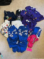 Full lot of assorted Disney and Looney Tunes clothing items laid out on floor, including tops, sweatshirts, and baby onesies in various colors and styles