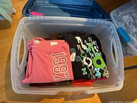 Open Rubbermaid bin showing folded assorted hoodies and sweatpants, pink sweatpants with 1987 patch and a black hoodie with graphic.